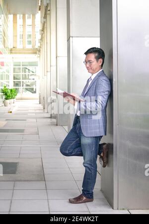 Uomo d’affari asiatico appoggiato contro la costruzione di pilastri, guardando il suo diario Foto Stock