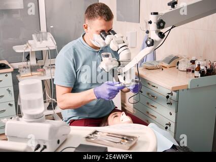 Dentista maschile che usa il microscopio dentale durante il controllo dei denti del paziente femminile. Donna sdraiata in sedia dentale mentre la stomatologa esamina i suoi denti. Concetto di odontoiatria, stomatologia e cura dentale. Foto Stock