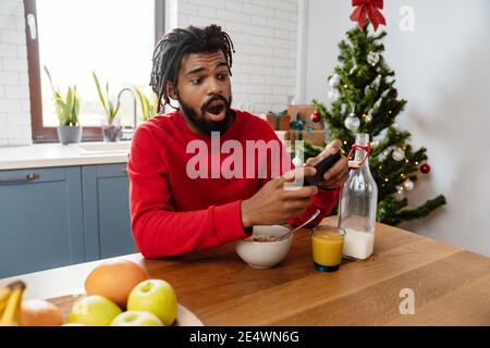 Uomo afro-americano scioccato che usa il telefono cellulare mentre fa colazione in una cucina accogliente Foto Stock