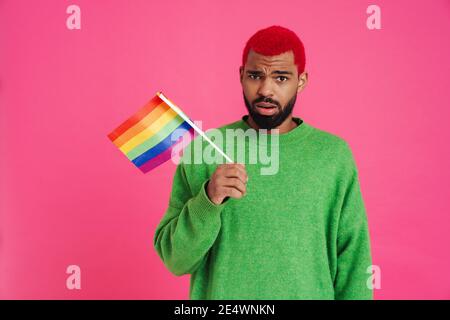 Confuso ragazzo afroamericano in posa con bandiera arcobaleno isolato sopra sfondo rosa Foto Stock