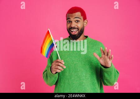 Gioioso ragazzo afroamericano che agita la mano e tiene la bandiera dell'arcobaleno isolato su sfondo rosa Foto Stock