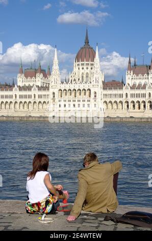 Budapest, Ungheria - 04 aprile 2012: Una giovane coppia siede sul lungofiume del Danubio Blu di fronte al monumentale Parlamento ungherese in Foto Stock