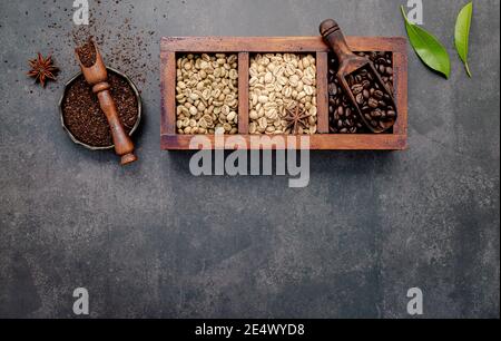 Chicchi di caffè verdi e marroni, tostati e scuri, in scatola di legno con sessole impostate su fondo di cemento scuro. Foto Stock