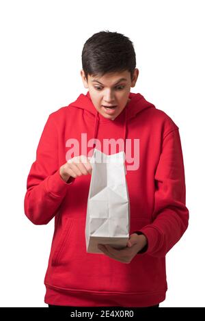 Sorprendo il ragazzo teenage troppo felice apertura borsa di carta bianca che guarda dentro pacchetto con espressione facciale Foto Stock