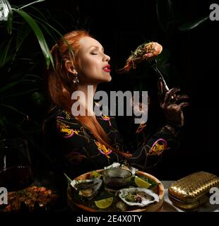Una ricca donna dai capelli rossi che mangia nel ristorante di pesce tiene un gamberetto a forcella o una langoutine. Tavolo da pranzo al tavolo che serve ostriche fresche Foto Stock