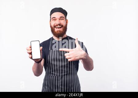 Ritratto dell'uomo dello chef felice in uniforme che punta sullo schermo dello smartphone su sfondo bianco. Foto Stock