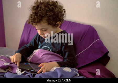 Dolce ragazzo di 4-5 anni con capelli ricci a letto sveglio al mattino, guardando cartoni animati su tablet, divertendosi in interni. Nuovo concetto normale Foto Stock