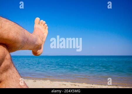 Le gambe incrociate dell'uomo si prendono il sole distendendo spensierato sulla sabbia accanto alla costa, sulla spiaggia pubblica. Foto Stock