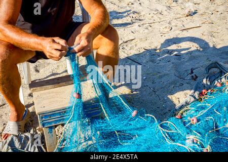 Un pescatore si siede su una sedia alla spiaggia, accumola la rete di pesca e la pulisce dalle conchiglie. Foto Stock