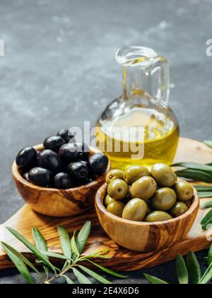 Set di olive verdi e nere e olio d'oliva su sfondo grigio. Diversi tipi di olive in ciotole di legno d'oliva e olio d'oliva su tagliere di legno e foglie d'oliva fresche. Spazio di copia. Verticale Foto Stock