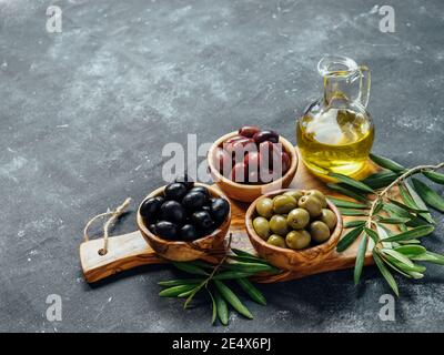 Set di olive verdi, nere e rosse o rosa e olio d'oliva su sfondo grigio. Diversi tipi di olive in ciotole di legno d'oliva e olio d'oliva su tagliere di legno. Spazio di copia Foto Stock