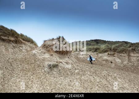 Un surfista che porta la sua tavola da surf e cammina sul sistema di dune di sabbia a Crantock Beach a Newquay in Cornovaglia. Foto Stock