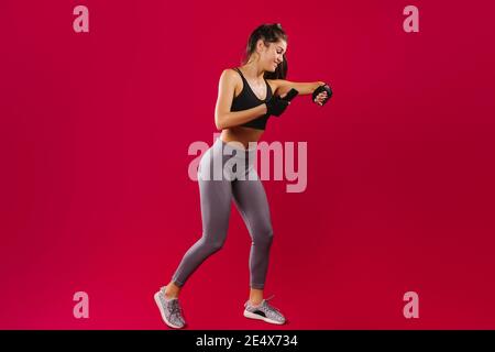 Fitness ragazza in abbigliamento sportivo durante l'allenamento osserva i suoi risultati su un braccialetto fitness e smartphone. Sfondo rosso e spazio vuoto dell'annuncio. Foto Stock