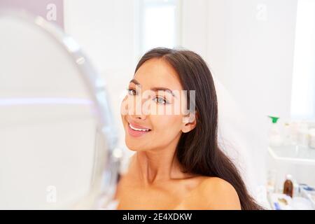 Sorridente giovane donna guarda il suo volto nello specchio dopo chirurgia plastica Foto Stock