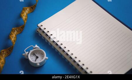 Vista su forchetta e coltello da cucina con misurazione a nastro e sveglia bianca. Spazio di copia carta bianca per notebook. Il concetto di digiuno intermittente guarisce Foto Stock