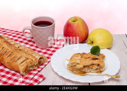 Apple roll servito. Un rotolo di strudel di mele fresco cotto in casa con una tazza di tè, una fetta appetitosa di torta di mele sul piatto e mele fresche su un Foto Stock