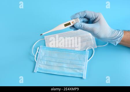 I guanti a mano contengono un termometro digitale contro le maschere mediche e lo sfondo blu. Concetto sanitario e medico. Foto Stock