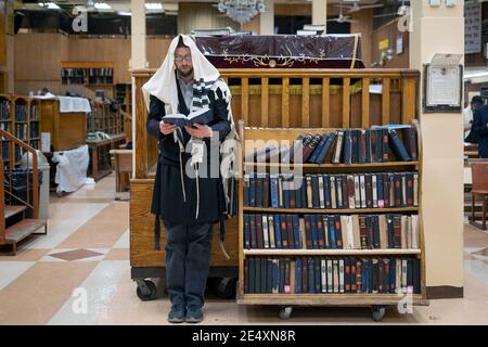 Un uomo ebreo ortodosso prega in una sinagoga mentre si trova da solo accanto a mensole di libri di preghiera. In una sinagoga vicina vuota a Brooklyn, New York. Foto Stock