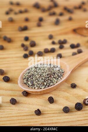 Macinato pepe nero su cucchiaio di legno con molto peppercorn sparsi su sfondo di legno Foto Stock