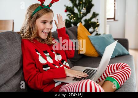 Felice bella ragazza che indossa corna di cervo giocattolo che agita mano e. uso del computer portatile mentre si è seduti sul divano di casa Foto Stock