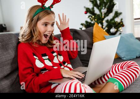 Felice bella ragazza che indossa corna di cervo giocattolo che agita mano e. uso del computer portatile mentre si è seduti sul divano di casa Foto Stock