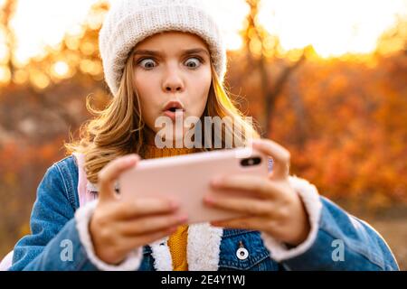 Immagine di giovane ragazza scioccata all'aperto in autunno parco utilizzando telefono cellulare Foto Stock