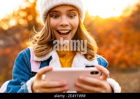 Foto di giovane ragazza felice scioccata all'aperto nel parco autunnale utilizzo del telefono cellulare Foto Stock