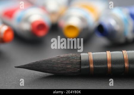 Pennello artistico per pittura ad acquerello primo piano su foglio di carta nera di album. Tubi di vernici in diversi colori su sfondo. Foto Stock