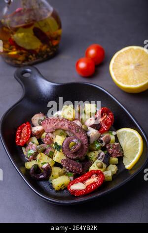 Insalata calda con polpo, patate, pomodori secchi, capperi e limone in una piccola padella nera. Primo piano, sfondo nero. Foto Stock