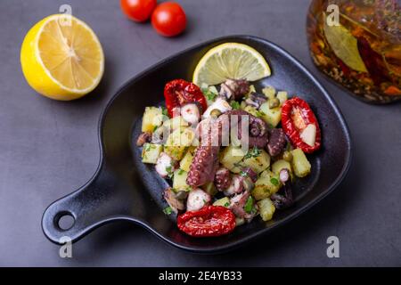 Insalata calda con polpo, patate, pomodori secchi, capperi e limone in una piccola padella nera. Primo piano, sfondo nero. Foto Stock