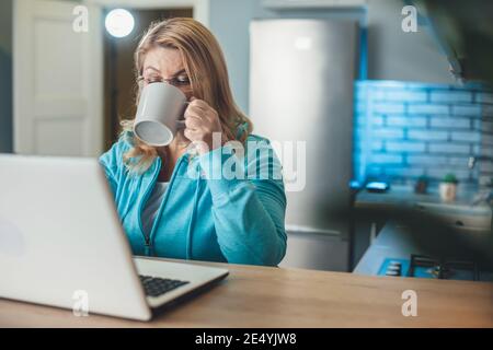 La donna bionda anziana impegnata sta bevendo una tazza di tè mentre si lavora da casa al computer portatile in cucina Foto Stock