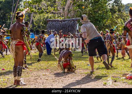 Un passeggero della nave da spedizione True North, indossando una camicia bianca e pantaloni blu, lancia la palla arancione durante una partita di cricket in stile Isole Trobriand a Kwebwaga, Isole Trobriand, Papua Nuova Guinea, un ballerino in costume accovacciato davanti al wicket, uomini in costume che danzano intorno a loro. Foto Stock