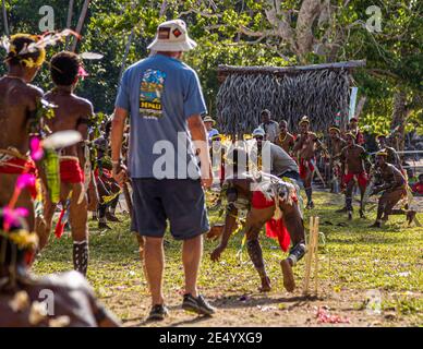Un passeggero della nave da spedizione True North si trova con un bastone di legno in mano davanti al wicket-keeper accovacciato durante una partita di cricket in stile delle Isole Trobriand a Kwebwaga, Isole Trobriand, Papua Nuova Guinea, ballerini in costume osservano la scena sullo sfondo. Foto Stock
