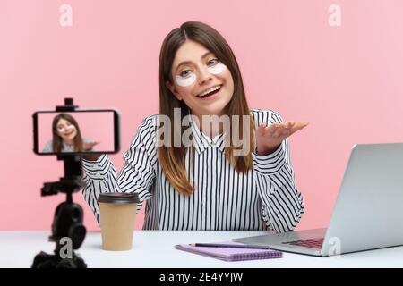 Positivo beauty blogger donna attivamente gesticulating registrazione video su fotocamera smartphone, chatting con seguaci, seduta con gli occhi patch sul viso. Foto Stock