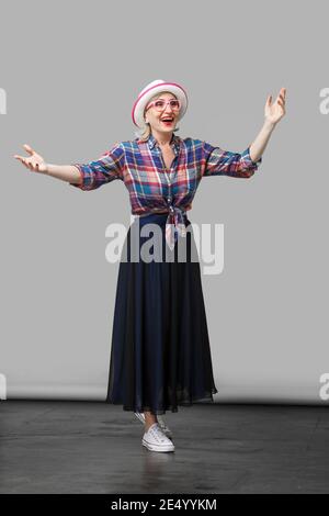 A piena lunghezza Ritratto di felice moderno ed elegante donna matura in stile casual con cappello e occhiali in piedi con le braccia sollevate e sorpresi di fronte alla ricerca Foto Stock