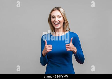 Buon lavoro! Ritratto di donna estremamente felice in abito blu stretto mostrando pollici e sorridendo ampiamente alla macchina fotografica, emozionato espressione. Indoor Foto Stock