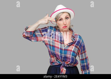 Ritratto di crazy moderno ed elegante donna matura in stile casual con cappello bianco in piedi e guardando la fotocamera con pistola arma gesto. piscina studio Foto Stock