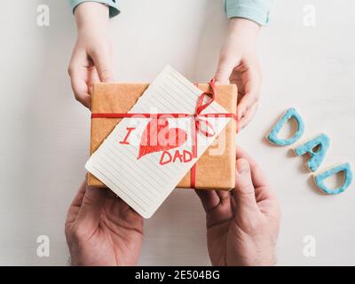 Bambino con le mani in mano e di uomo adulto le mani, bella confezione regalo, nastro e i cookie di vetrate su un bianco, sullo sfondo di legno. Vista superiore, close-up. Preparazione per TH Foto Stock