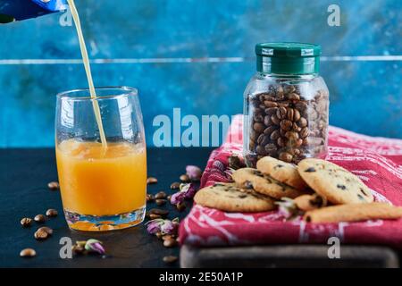 Biscotti al cioccolato, chicchi di caffè in un recipiente di vetro e un bicchiere di succo d'arancia su un tavolo scuro Foto Stock