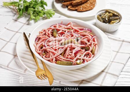 Insalata di salsiccia tedesca di salsiccia di lyoner a fette con cetrioli e. cipolla rossa servita su un piatto bianco con posate dorate e pane di segale su un legno bianco Foto Stock