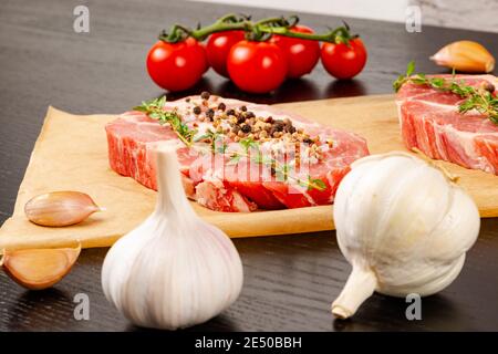 bistecche di maiale crude tagliate al momento con spezie e timo su un foglio di pergamena. accanto ai pomodori rossi maturi e all'aglio Foto Stock