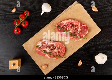 bistecche di maiale crude tagliate al momento con spezie e timo su un foglio di pergamena. accanto ai pomodori rossi maturi e all'aglio Foto Stock