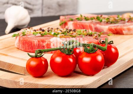 bistecche di maiale crudo appena tritate con spezie e timo su un tagliere. accanto ai pomodori rossi maturi e all'aglio Foto Stock