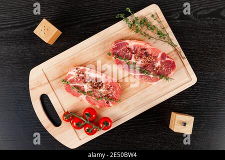 bistecche fresche di maiale crudo tritate con spezie, pomodori e timo su un tagliere di cucina su un tavolo di legno nero. accanto allo shaker per pepe e al sale shake Foto Stock