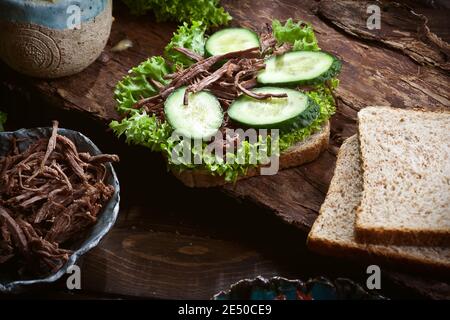 assemblaggio del sandwich di carne con foglie di lattuga verde, pomodori secchi, cetrioli freschi e pane di segale o di grano, su sfondo di legno rustico scuro, nobo Foto Stock