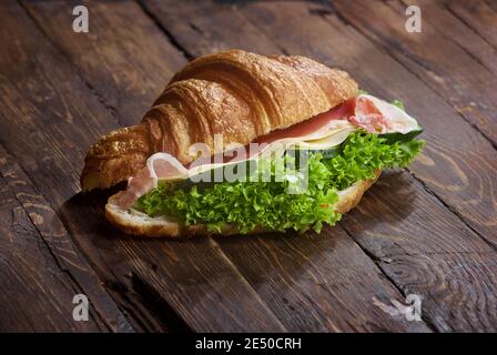 un panino di croissant con fette di carne di jamon, foglie di insalata di lattuga verde, cetrioli freschi e formaggio, su una rustica superficie di tavola in legno, copyspace Foto Stock