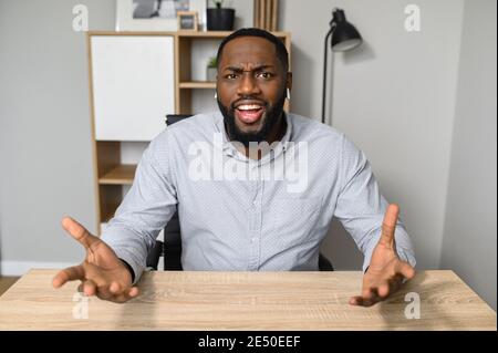 Un giovane e bell'uomo d'affari afro-americano è seduto alla scrivania e punta alla macchina fotografica, la sensazione di confusione, il proprietario di affari di successo è Foto Stock