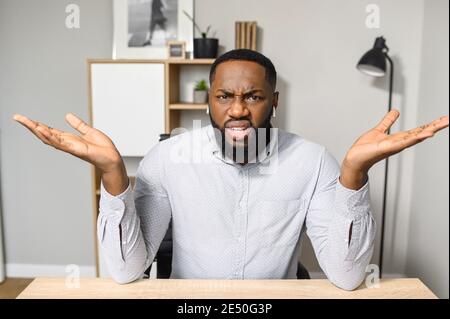 Un giovane e bell'uomo d'affari afro-americano è seduto alla scrivania e alzando le mani in su, sentendosi confuso, proprietario riuscito di affari è Foto Stock