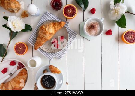 Disposizione piatta di un tavolo per la colazione con croissant con marmellata, caffè, lamponi, arancio sangue, uova sode e fiori su un tavolo di legno bianco, orizzontale Foto Stock