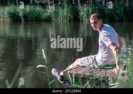 Felice uomo rilassato seduto sul molo di legno su un lago godendo di una soleggiata giornata estiva circondato da verde e alberi guardando la macchina fotografica. Salute mentale, rebo Foto Stock